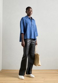 Blaues Oversize-Denimhemd mit Kragen, vorne mit Knopfleiste, kombiniert mit schwarzen Lederhosen und weißen Sneakern, während eine hellbraune Tragetasche gehalten wird.