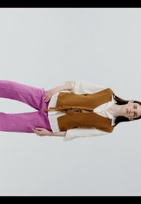 Femme portant un chemisier blanc, un gilet en maille sans manches marron avec ceinture, et un pantalon violet vif, debout devant un fond clair uni.