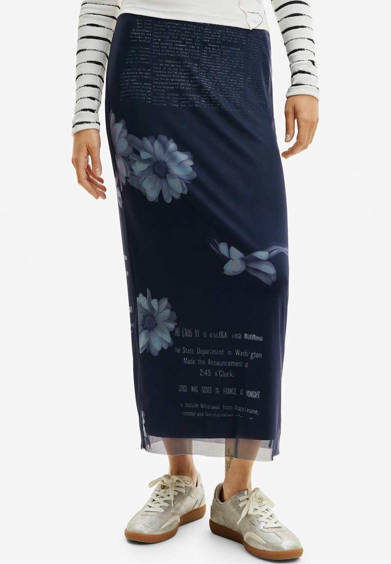 Femme portant une jupe midi bleu marine à imprimé floral et texte, un haut à manches longues rayé, et des baskets beiges, debout devant un fond blanc.