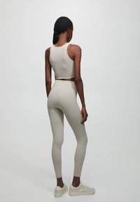 Donna con capelli intrecciati, che indossa un top corto senza maniche grigio chiaro, leggings a vita alta abbinati e sneakers bianche, in piedi su uno sfondo bianco.