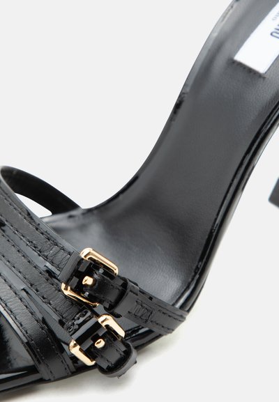 Μαύρο σανδάλι με ψηλό τακούνι από δέρμα πατέντας, με δύο χρυσού χρώματος αγκράφες μπροστά και κομψό, κυρτό πέλμα.