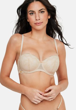 Femme aux longs cheveux bruns portant un soutien-gorge balconnet en dentelle beige et une culotte assortie, debout devant un fond blanc, les mains légèrement jointes.