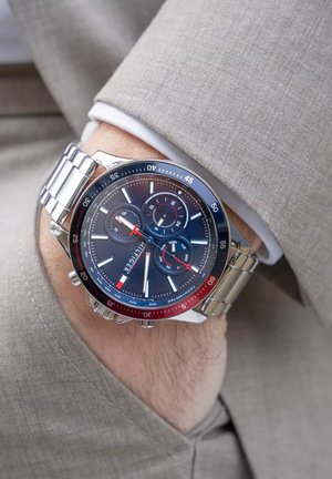 Pols van een man met een zilveren Tommy Hilfiger chronograafhorloge dat de datum en dag aangeeft, gedragen onder een beige colbert en een witte overhemdmanchet.