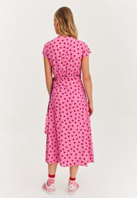 Růžové šaty s puntíky, krátkými rukávy, zúženým pasem a midi délkou; uvázané na boku; nošené s růžovými teniskami a červenými pruhovanými ponožkami.