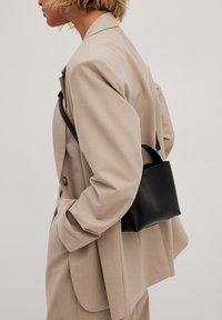 Schwarze Lederhandtasche mit strukturiertem Design und einem oberen Griff, getragen zu einem beige maßgeschneiderten Blazer, der eine glatte Textur und minimalistisches Design zeigt.