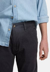 Camisa de manga corta de color azul claro abotonada sobre pantalones negros con un botón visible y detalles de bolsillo, que muestra una tela suave y texturizada.