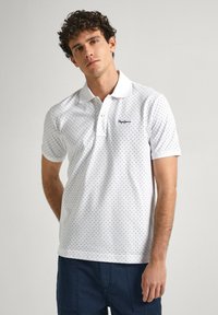 Biele polo tričko s námorníckym modrým trojuholníkovým vzorom, krátke rukávy a zapínanie na gombíky. Logo značky na ľavej strane hrude.
