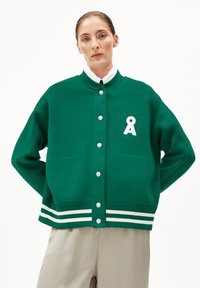 Veste en tricot verte avec un col rond, des boutonnières blanches, deux poches avant et des accents rayés au niveau de l'ourlet. Présente un logo blanc.