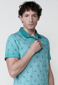 Téglazöld, rövid ujjú pólóing apró levél- és teknős mintákkal, texturált felülettel. Kontrasztos gallérral és gombokkal rendelkezik.