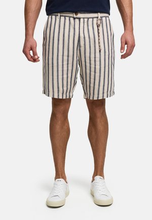 Mężczyzna ubrany w beżowe szorty z pionowymi granatowymi paskami i białe trampki, stojący na neutralnym tle.