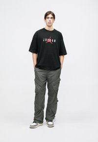 Crna majica kratkih rukava s "JORDAN" logotipom u crvenoj boji, uparena s sivim cargo hlačama koje imaju više džepova i patentnih zatvarača. Crno-bijele tenisice.