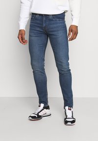 Skinny denim jeans in dark blue with subtle fading and whiskering. Paired with black and white sneakers featuring red accents.