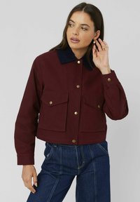 Veste courte bordeaux avec un col en velours côtelé, deux poches avant et des boutons dorés. Associée à un jean taille haute bleu.