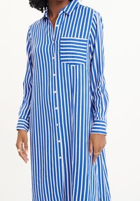 Žena oblečená v modro-bílých svisle pruhovaných košilových šatech s přední kapsou a dlouhými rukávy, ukazuje ruce s prsteny.