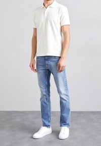 Ljusbeige pikétröja med korta ärmar, kombinerad med ljusblå jeans och vita sneakers. Enkelt design utan synliga mönster eller detaljer.