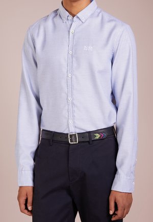 Mand iført lyseblå mønstret skjorte med knapper, sorte bukser og sort bælte med farverige pil-designs nær spænde.