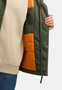 Grøn ydre jakke med orange quiltet for, der har en frontlynlås, sidelåse og et beige indre tøj, der er synligt i bunden.