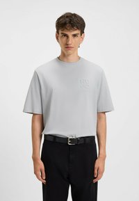 Lysegrå kortærmet t-shirt med rund halsudskæring og prægede "HUGO" logo. Båret med sorte bukser og et sort bælte med en sølvspænde.