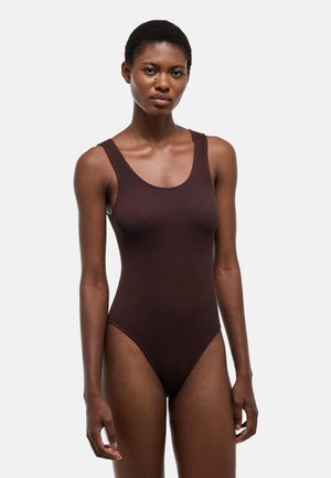 Femme portant un maillot de bain une pièce sans manches, côtelé, de couleur brun foncé, debout devant un fond clair uni.