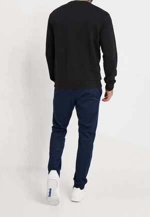 Man die een zwart shirt met lange mouwen, marineblauwe broek en witte sneakers draagt en wegloopt tegen een effen witte achtergrond.