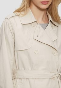 Beige Trenchcoat mit doppelter Knopfleiste, großen Knöpfen, weitem Kragen und gebundenem Taillenbereich. Hergestellt aus leichtem Material.