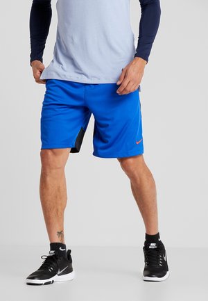 Hombre que lleva pantalones cortos deportivos azules, zapatillas de entrenamiento negras de Nike, calcetines negros y una camisa de manga larga azul claro, de pie contra un fondo liso.