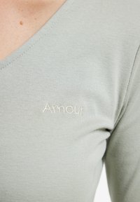 Light green, ribbed fabric with a V-neck design featuring "Amour" embroidered in white on the chest. Smooth texture and fitted shape.