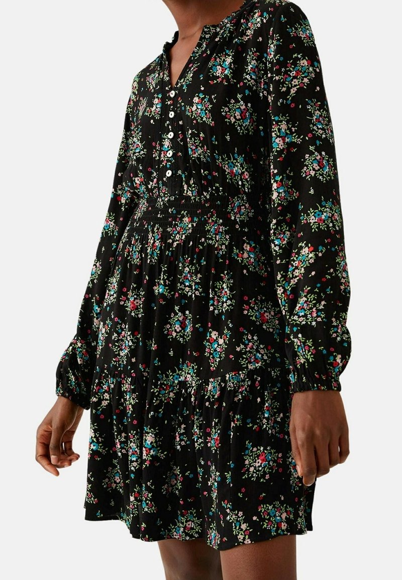 Femme portant une robe noire à manches longues avec un petit motif floral coloré et des boutons blancs sur le haut devant.