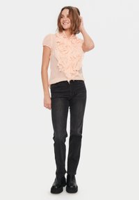 Blusa ruggente color pesca con maniche corte, caratterizzata da un motivo floreale testurizzato sul davanti e abbinata a jeans scuri e stivali neri.