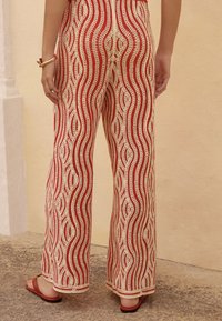 Rote und cremefarbene gemusterte weite Hosen mit wellenförmigen Linien, strukturiertem Stoff und einem lockeren Schnitt, kombiniert mit einfachen flachen Sandalen.