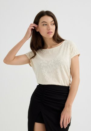 Top corto de color beige claro con una sutil textura floral, combinado con una falda negra de canalé con una abertura lateral y detalles fruncidos.
