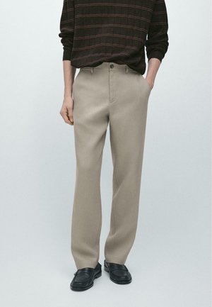 Uomo che indossa pantaloni beige a gamba dritta, mocassini neri e una camicia a manica lunga marrone scuro con sottili righe orizzontali, in piedi con le mani in tasca.