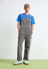 Gray denim overalls with adjustable straps, front pocket, and wide legs, paired with a blue t-shirt and light blue sneakers. Hat on head.
