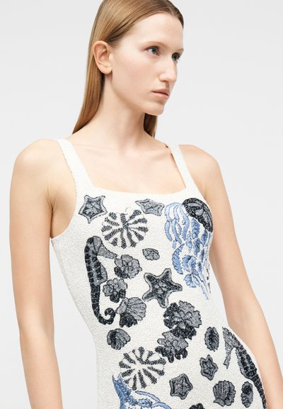 Γυναίκα που φοράει λευκό αμάνικο φόρεμα με κεντήματα μαύρης και μπλε θαλάσσιας ζωής, περιλαμβάνοντας ιππόκαμπους, αστερίες και κοράλλια.