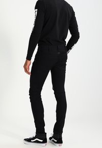 Černé džíny slim fit, s bočními kapsami a texturovanou látkou, spárované s černobílými teniskami. Viditelný černý top s dlouhým rukávem.