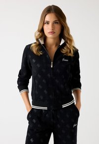 Zwarte zip-up jas met subtiel logopatroon, geribde manchetten en zoom, en een witte kraag. Gekleed met bijpassende zwarte broek.