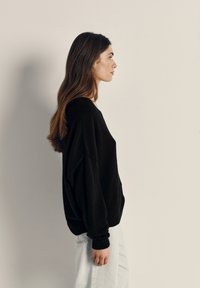 Maglione nero oversize con maglia testurizzata, caratterizzato da maniche larghe e una vestibilità rilassata. Indossato con jeans chiari e larghi.