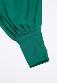 Tissu de blouse verte avec manches froncées, doté d'un poignet ajusté orné de trois boutons recouverts de tissu. Texture douce, finition lisse.