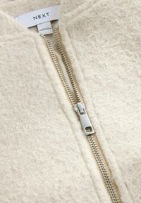 Jachetă crem cu fermoar, realizată dintr-un material texturat, moale, cu fermoar argintiu vizibil și detalii cu etichetă la guler.