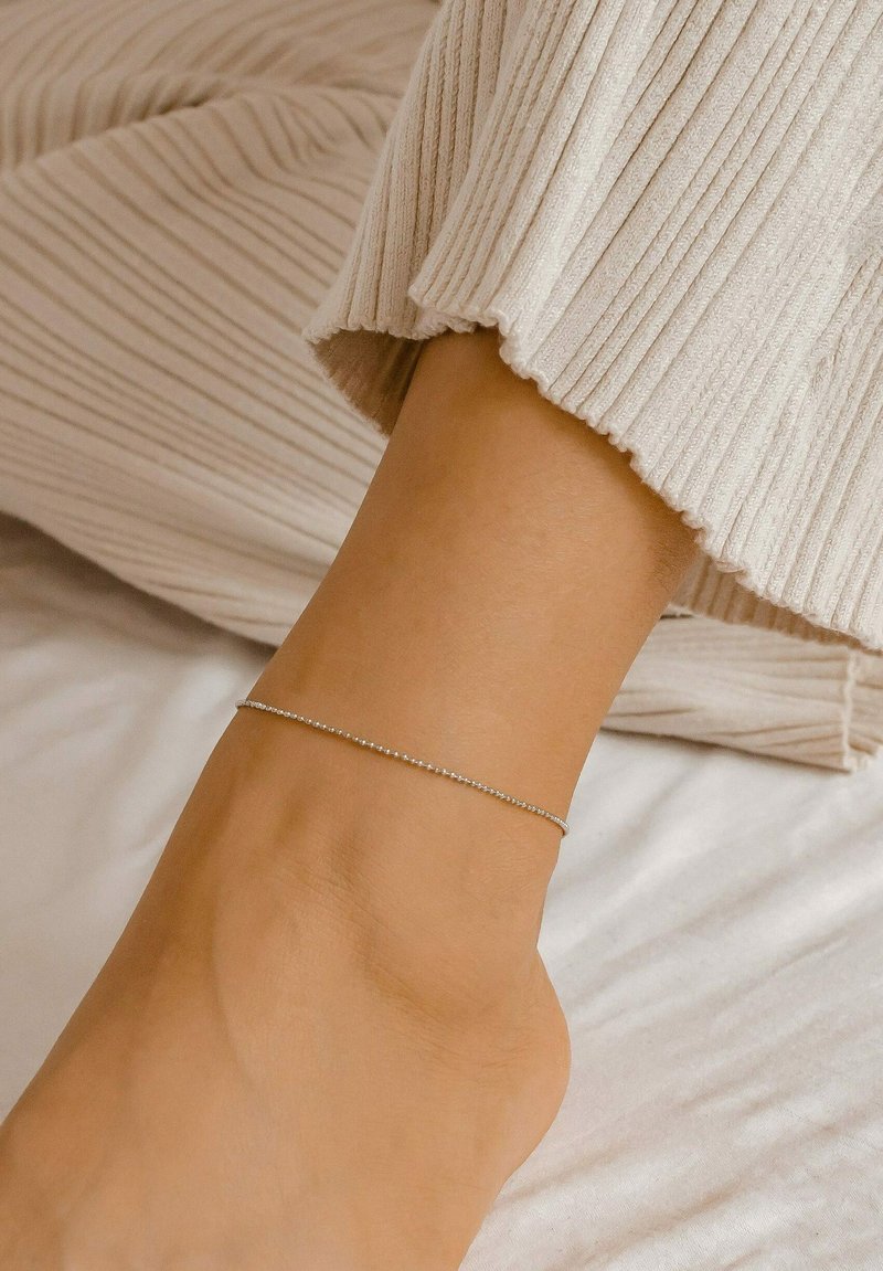 Silver ankle bracelet with a delicate chain design, featuring small round beads, worn on a bare ankle against a light-colored fabric background.