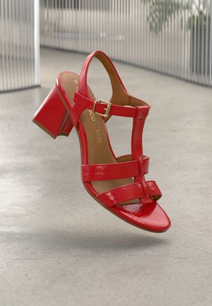 Red patent leather heeled sandals with adjustable ankle strap, open toe, and rectangular block heel. Features minimalistic strap design.