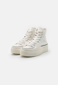 High-top sneakers in white canvas with rubber soles, featuring metal eyelets, flat cotton laces, and a black stripe on the midsole.