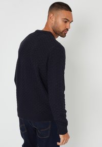 Marineblauer Pullover mit einem strukturierten, gemusterten Strickdesign, gerippten Bündchen und Saum. Getragen mit dunklen Jeans, die eine Seiten- und Rückansicht zeigen.