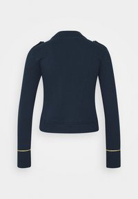 Veste cintrée bleu marine vue de dos, avec des manches longues et une fine bande dorée près de chaque poignet. Aucun bouton ou fermeture éclair visible.