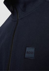 Námořnicky modrá mikina s kapucí na zip se malou čtvercovou nášivkou s nápisem "BOSS" na hrudi, ukazující strukturu látky a detaily zipu.