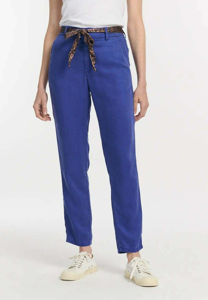 Helderblauwe chinos met een relaxte pasvorm, voorzien van een bruin luipaardprintriem, zijzakken en casual witte sneakers. Glad stoftextuur.