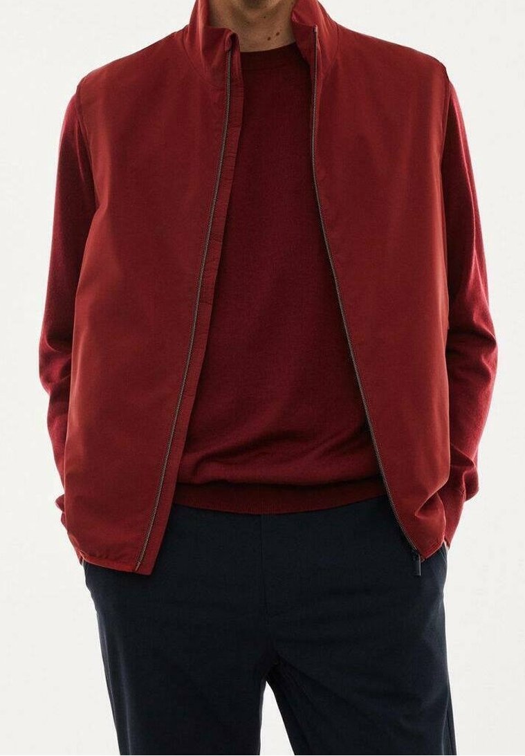 Homme portant une veste zippée rouge foncé sur un pull rouge assorti avec un pantalon foncé, les mains dans les poches, sur un fond blanc uni.