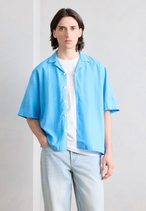 Junger Mann mit schulterlangem dunklem Haar, der ein hellblaues, kurzärmeliges Hemd über einem weißen T-Shirt und hellblauen Jeans trägt.