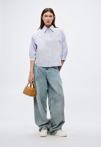 Ανοιχτό μπλε πουκάμισο με κοντά μανίκια και γιακά, σε συνδυασμό με φαρδιά τζιν σε ανοιχτό χρώμα. Κρατάει μια μικρή ταμπά τσάντα χειρός και φοράει λευκά αθλητικά παπούτσια.