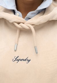 Felpa in corduroy beige con tasca frontale, logo "Superdry" ricamato e punte dei lacci grigie. Camicia color blu chiaro con colletto sottostante.